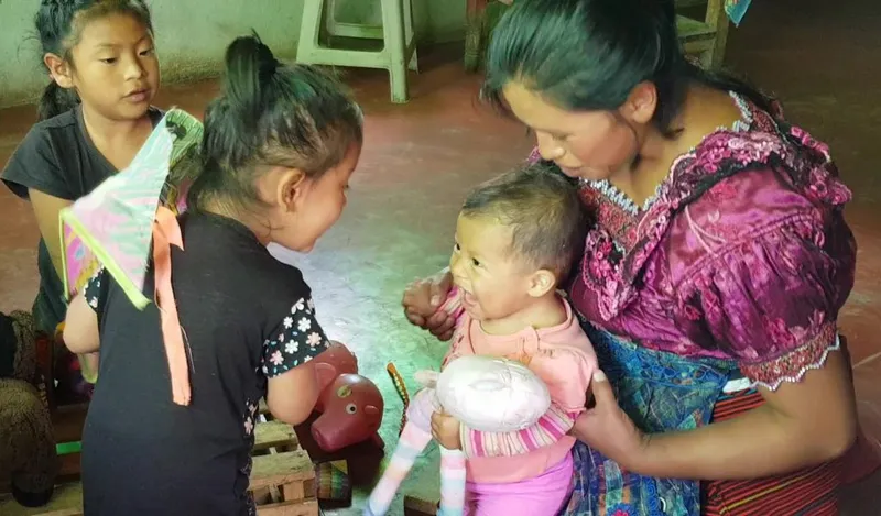 A mother holds her baby while older children play nearby during a child development visit.