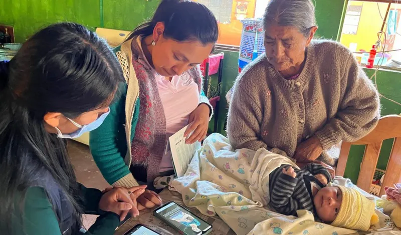 Two health workers review an infant development app on a smartphone with a mother and grandmother beside a swaddled newborn.