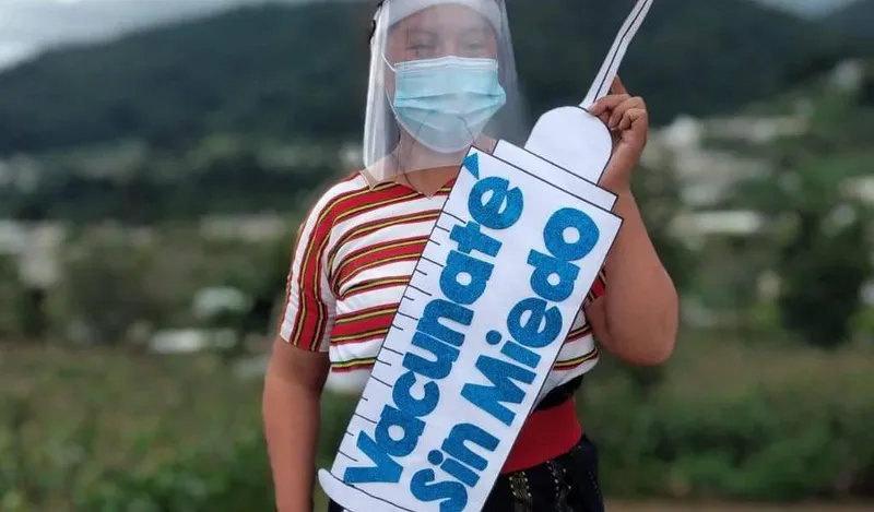 A young community member in a mask and face shield holds a large cut-out syringe with the Spanish slogan “Vacúnate sin miedo” (Get vaccinated without fear).
